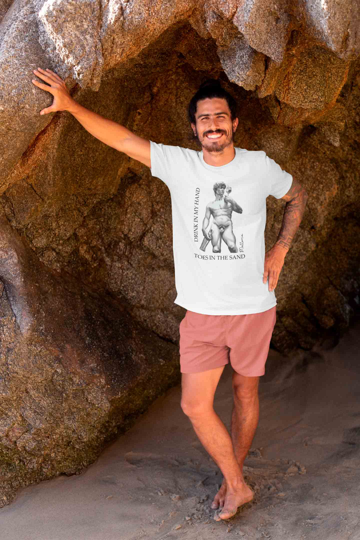 Dunkelhaariger muskulöser Mann lehnt lässig an einem Holzpfahlzaun und blickt nach links. Er trägt ein weißes Shirt mit schwarzen Palmen und der Aufschrift "Mallorca...wir sehen uns bald!"